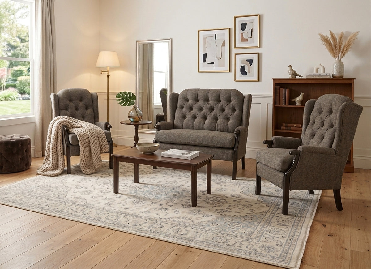 A styled living room featuring a gray tufted three-seat sofa, two matching gray wingback armchairs, a dark wood coffee table, a large patterned area rug, and a floor lamp, set against a warm neutral wall.