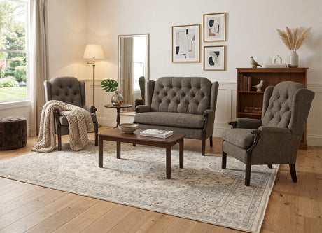 A styled living room featuring a gray tufted three-seat sofa, two matching gray wingback armchairs, a dark wood coffee table, a large patterned area rug, and a floor lamp, set against a warm neutral wall.
