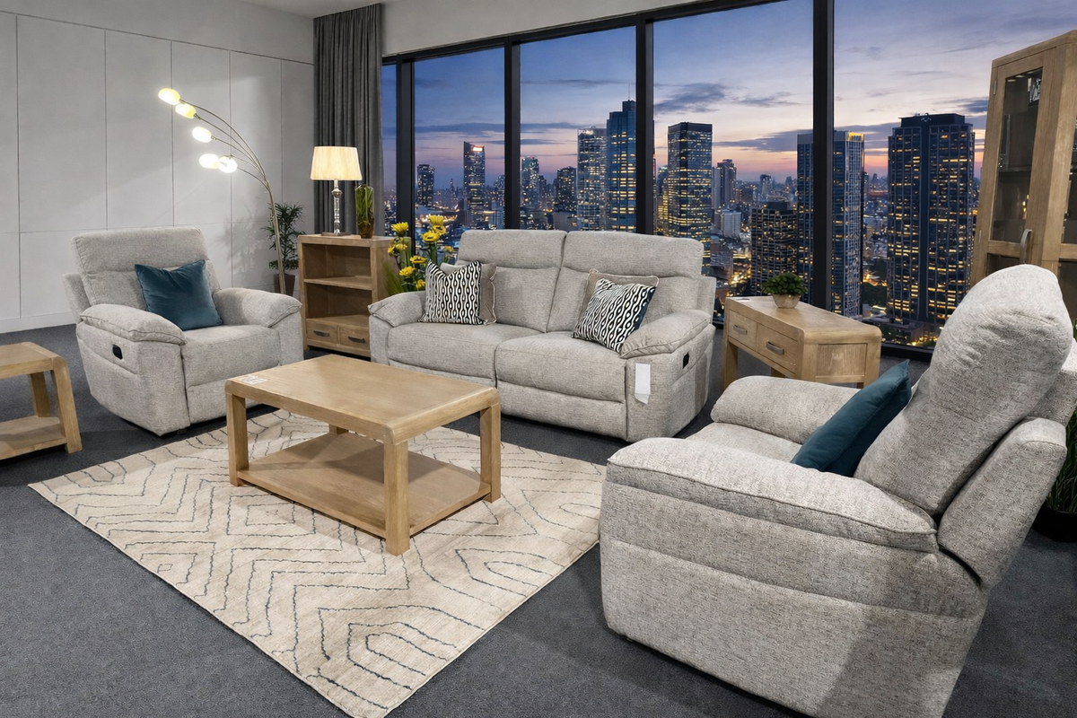 A staged living room featuring the Vera beige upholstered recliner, loveseat, and matching coffee table, arranged on a patterned rug in front of large windows showing a city skyline at dusk.