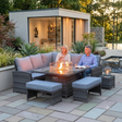 Outdoor amalfi patio conversation set with a dark woven wicker sectional sofa, two matching ottomans, a side table, and a glass-top fire pit table with visible flames, shown with a couple sitting and a modern pool house in the background at furniture store ni 