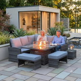 Outdoor amalfi patio conversation set with a dark woven wicker sectional sofa, two matching ottomans, a side table, and a glass-top fire pit table with visible flames, shown with a couple sitting and a modern pool house in the background at furniture store ni 