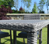 Gray wicker outdoor table and chairs in a garden setting with trees and stone wall in the background at the furniture store ni